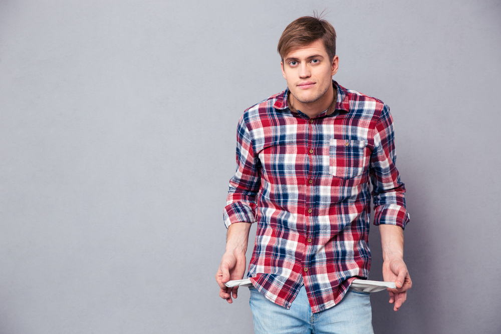 Poor handsome young man in checkered shirt and jeans showing empty pockets over grey background